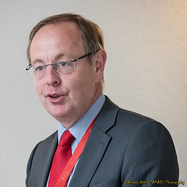 Patrick Child with a red tie in front of a plain background